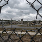 Clôture de base militaire avec barbelés et un drone visible dans le ciel nuageux