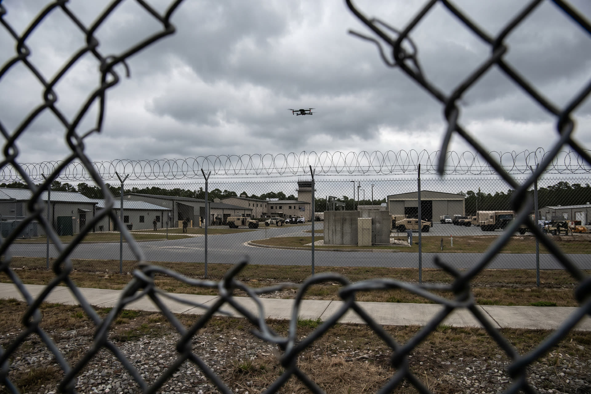 Clôture de base militaire avec barbelés et un drone visible dans le ciel nuageux